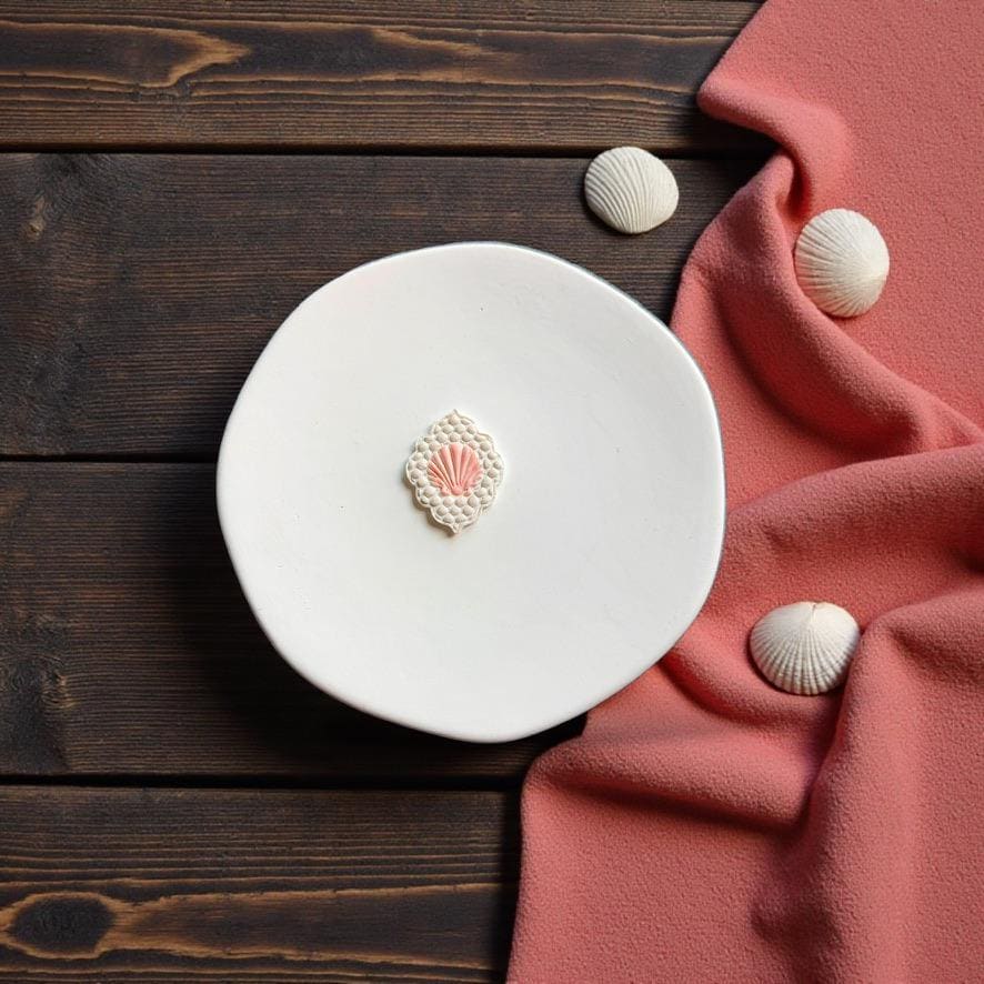 Finished lace seashell polymer clay earring displayed on white ceramic plate with coastal shell decor and pink fabric backdrop