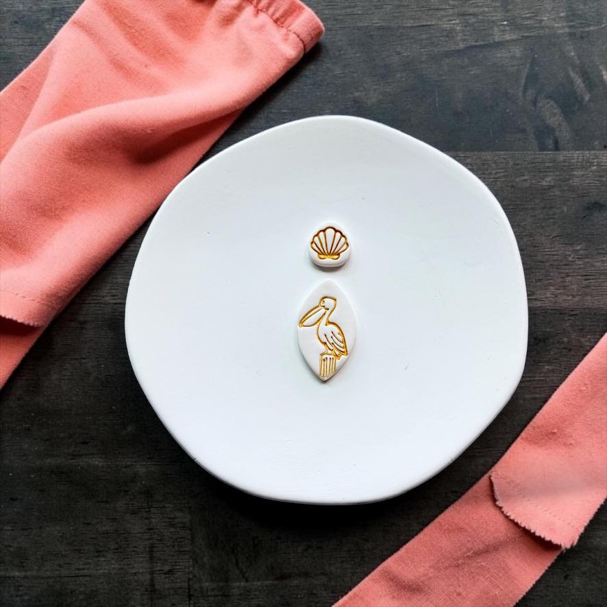 Finished pelican and seashell topper clay pieces displayed on a white ceramic plate with coral-colored fabric.