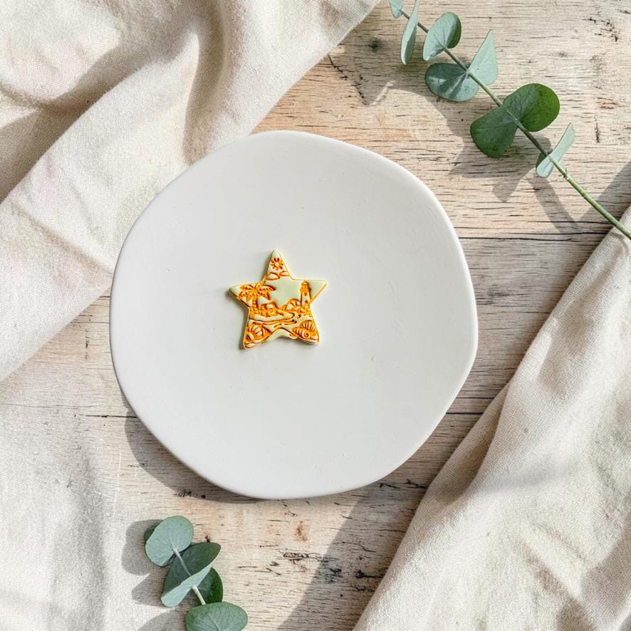 Finished star-shaped beach scene polymer clay earring on a white ceramic plate with eucalyptus accents.