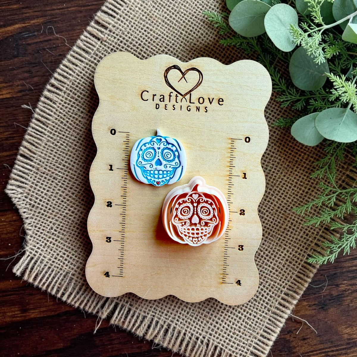 Finished sugar skull pumpkin earring beside the matching polymer clay cutter displayed on a Craft Love Designs size board.