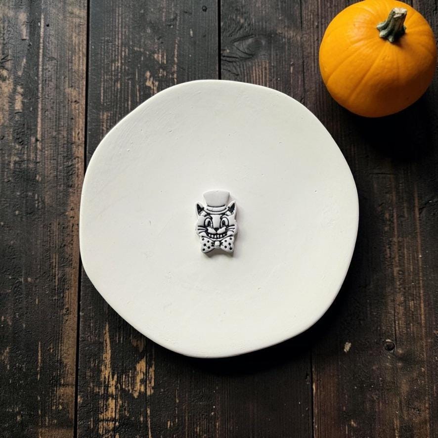 Finished white and black vintage Halloween cat with pointed ears, top hat, wide smiling face, whiskers, and oversized bow tie, centered on a white round surface with an orange pumpkin nearby.