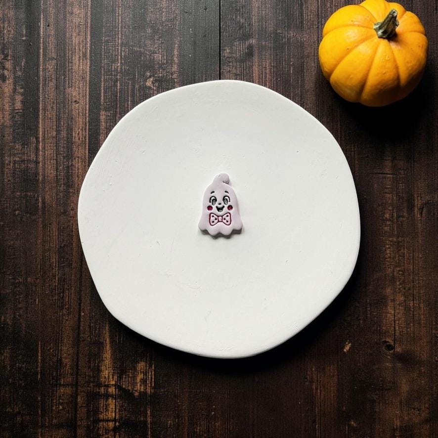 Finished vintage-style white ghost clay piece with expressive black facial details, rosy pink cheeks, and a pink bow tie, displayed on a white round surface with an orange pumpkin in the upper right.
