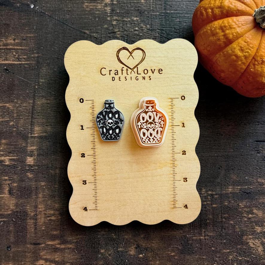 Black and gray finished skull potion bottle beside a larger peach-colored version on a Craft Love Designs wooden measuring board with an orange pumpkin in the background.