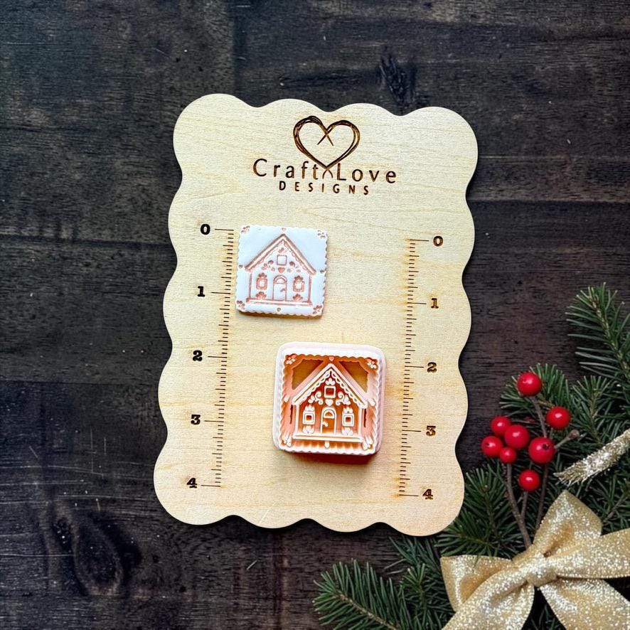 Two sizes of gingerbread house designs displayed on a Craft Love Designs wooden measuring board. A smaller white house with brown details appears above a larger brown and cream house, with Christmas greenery, red berries, and a gold bow beside the board.