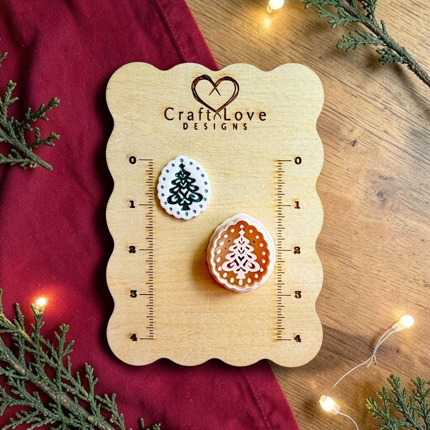 Finished white oval Christmas tree clay piece with a dark green decorative tree and dotted border beside the matching polymer clay cutter on a wooden Craft Love Designs sizing board with red fabric, evergreen branches, and holiday lights.