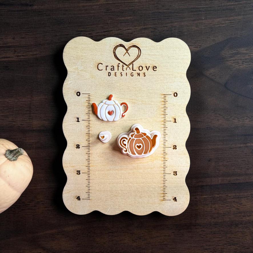 Finished pumpkin teapot clay piece and matching polymer clay cutter displayed on a wooden Craft Love Designs sizing board, with a small separate heart-shaped clay piece beside them and a pale pumpkin at the edge of the photo.