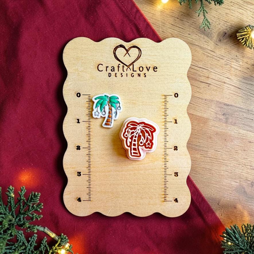 Finished Christmas palm tree clay piece with green palm leaves, brown trunk, and hanging silver ornaments beside the matching polymer clay cutter on a wooden Craft Love Designs sizing board with red fabric, greenery, and holiday lights.