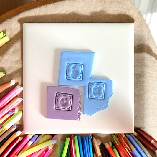 Two square silicone molds with letter stamps on a white surface, surrounded by colorful pens.