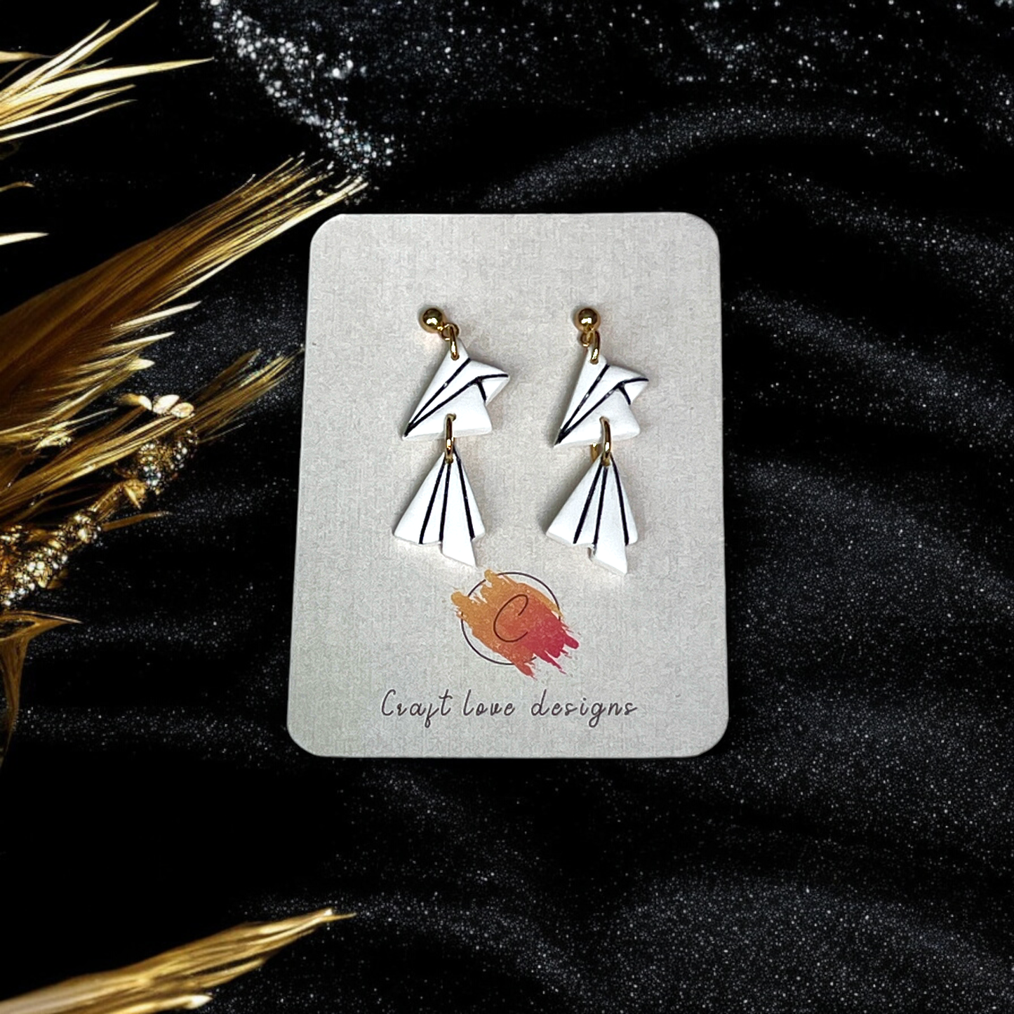 White geometric dangle earrings with angular design and black line detailing, displayed on a card against a black, textured backdrop with golden accents. 