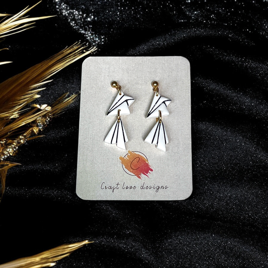 White geometric dangle earrings with angular design and black line detailing, displayed on a card against a black, textured backdrop with golden accents. 