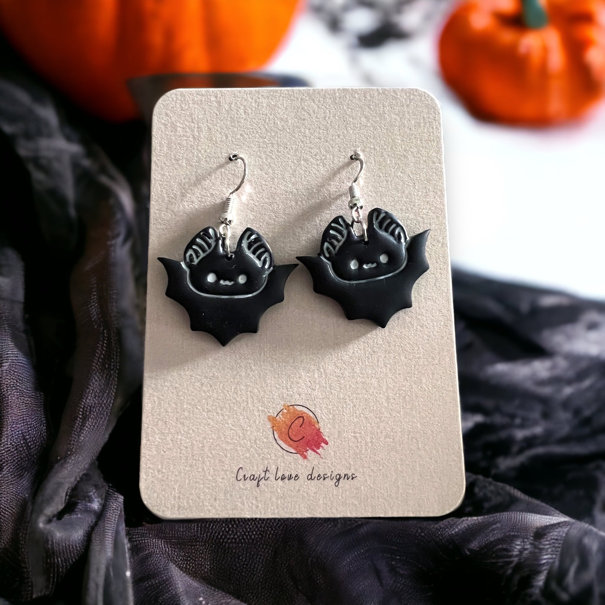 Pair of black bat-shaped dangle earrings with cute detailing, displayed on a beige card with festive Halloween pumpkins in the background.