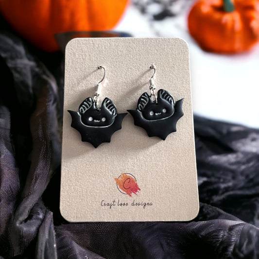 Pair of black bat-shaped dangle earrings with cute detailing, displayed on a beige card with festive Halloween pumpkins in the background.