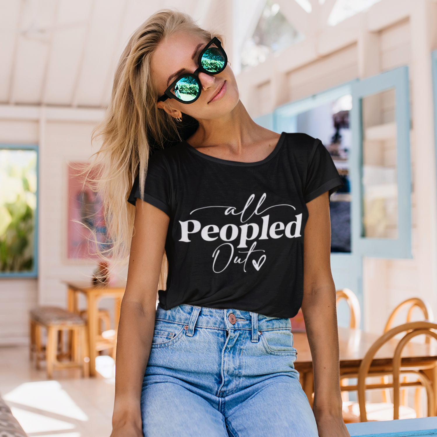Stylish woman wearing a black "All Peopled Out" t-shirt with sunglasses and high-waisted jeans, enjoying a relaxed moment in a sunny, cozy café.