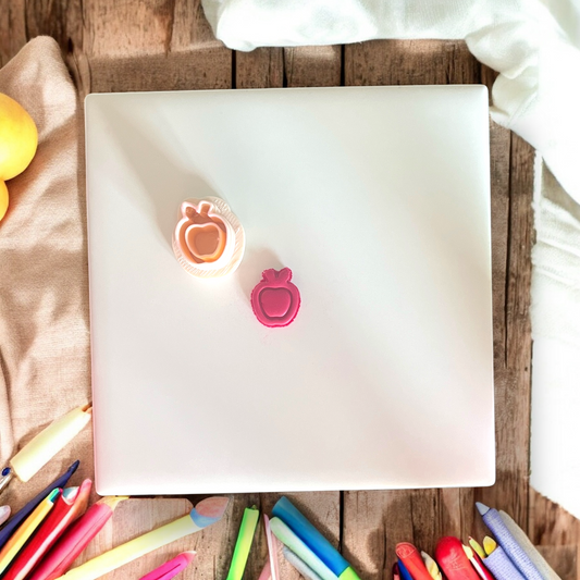Orange apple cutter and pink clay cutout side by side on a flat tile with scattered pencils.