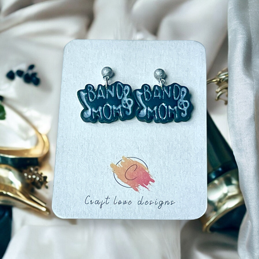 "Band Mom" drop earrings with treble clef design, attached to sparkly stud backs, displayed on a jewelry card over elegant white fabric. 