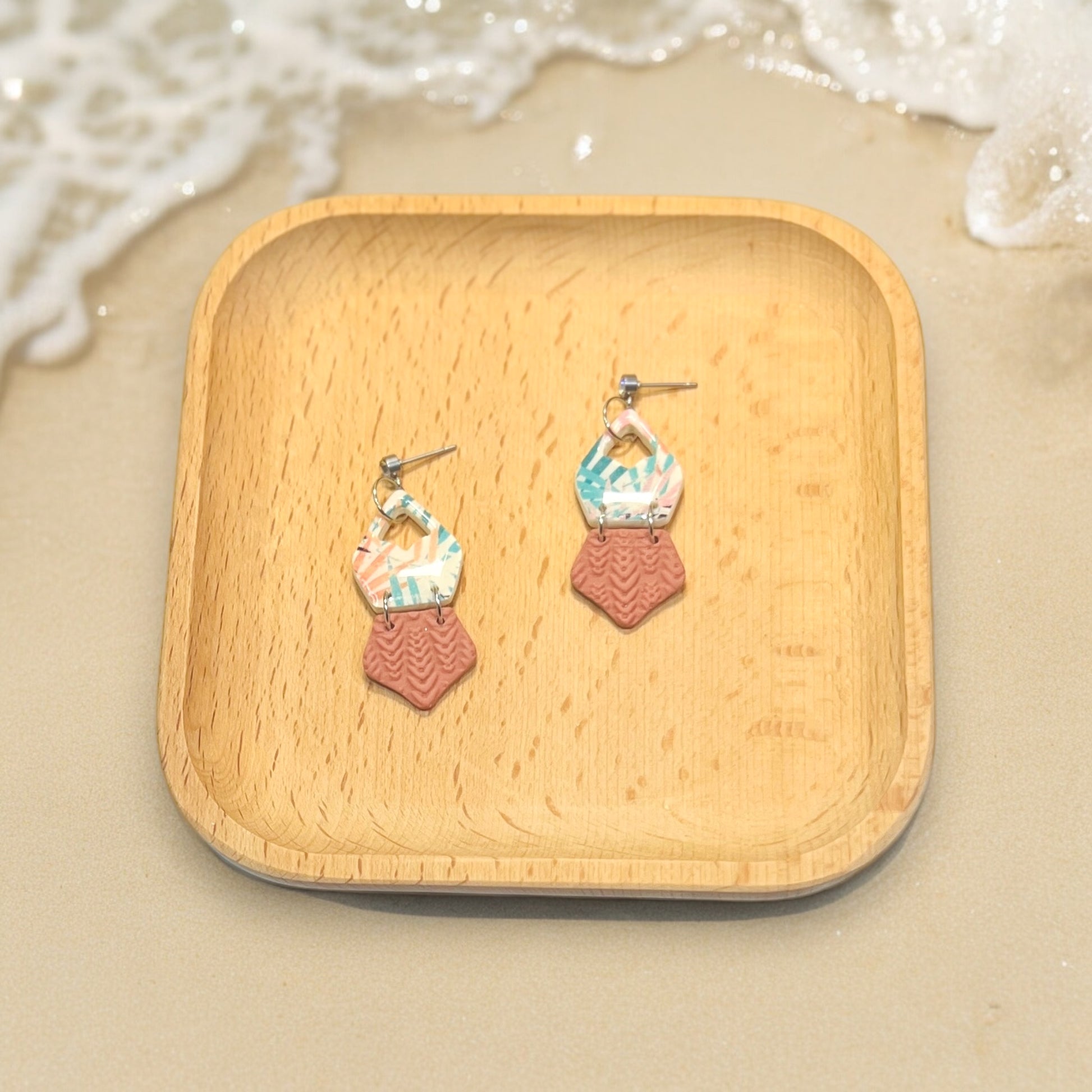 Multicolor design polymer clay dangle earrings presented on a wooden tray with beach sand and seashells in the background.
