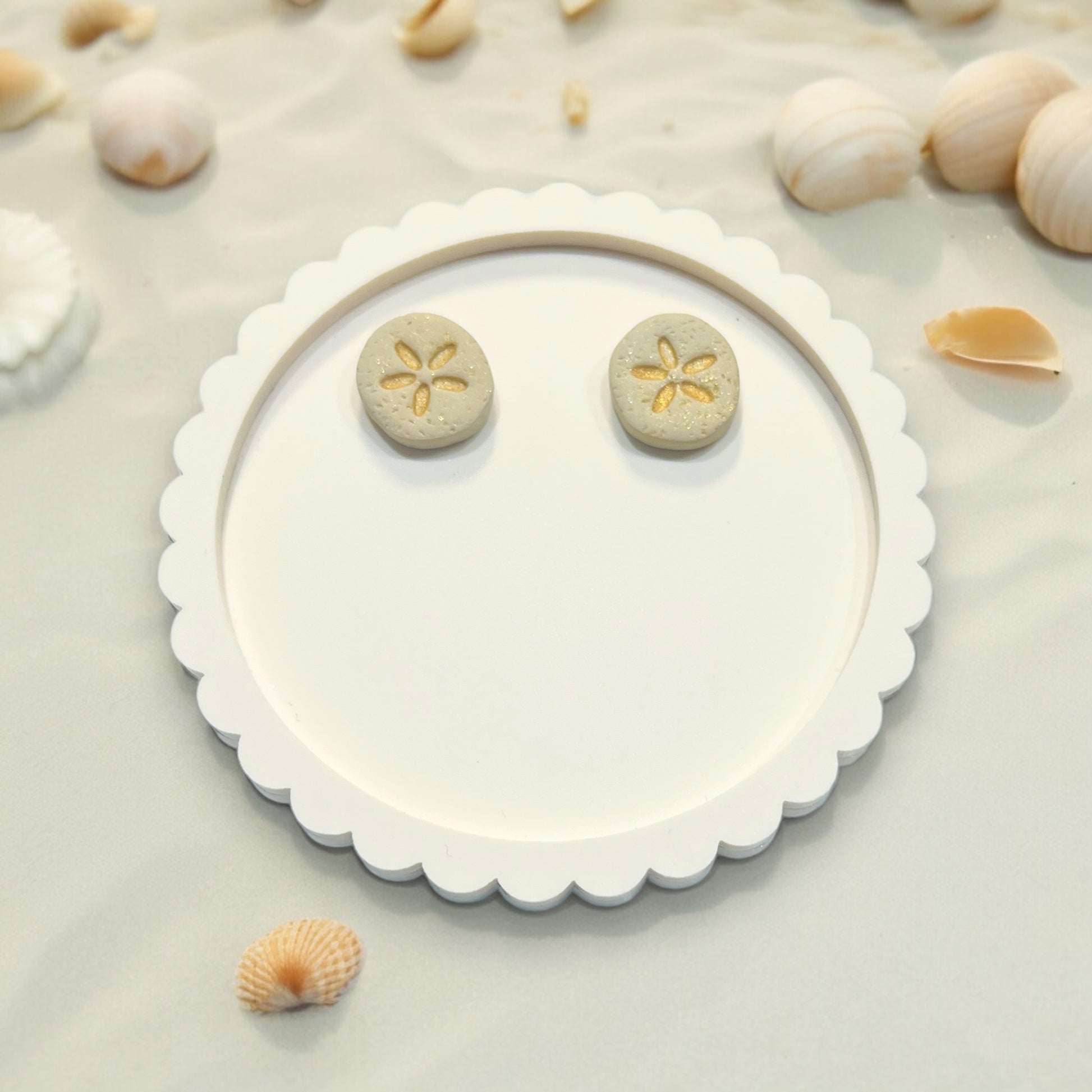Coastal-inspired sand dollar polymer clay earrings placed on a decorative tray surrounded by soft beige sand and shells. 