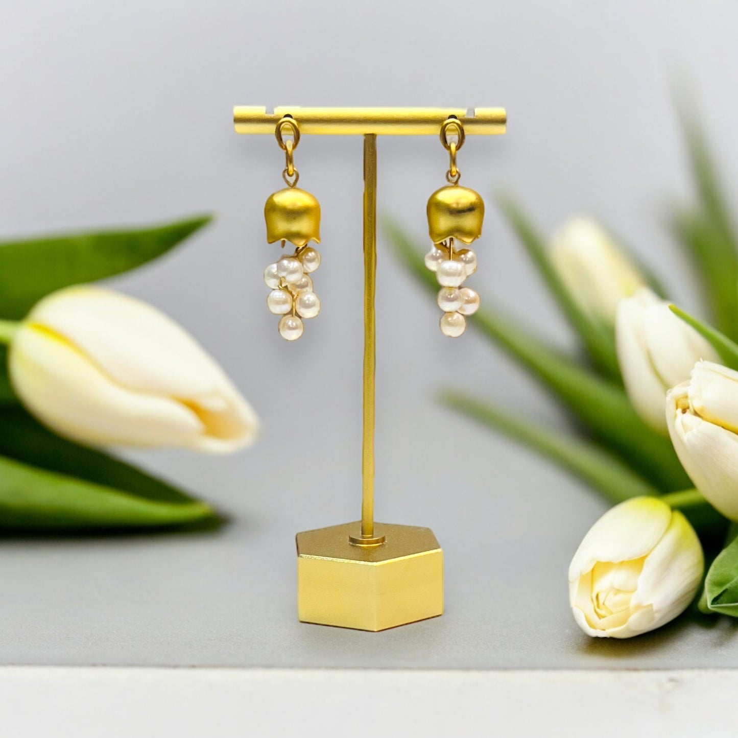 Gold bellflower earrings adorned with delicate pearls, displayed on a minimal golden earring stand next to fresh white tulips.