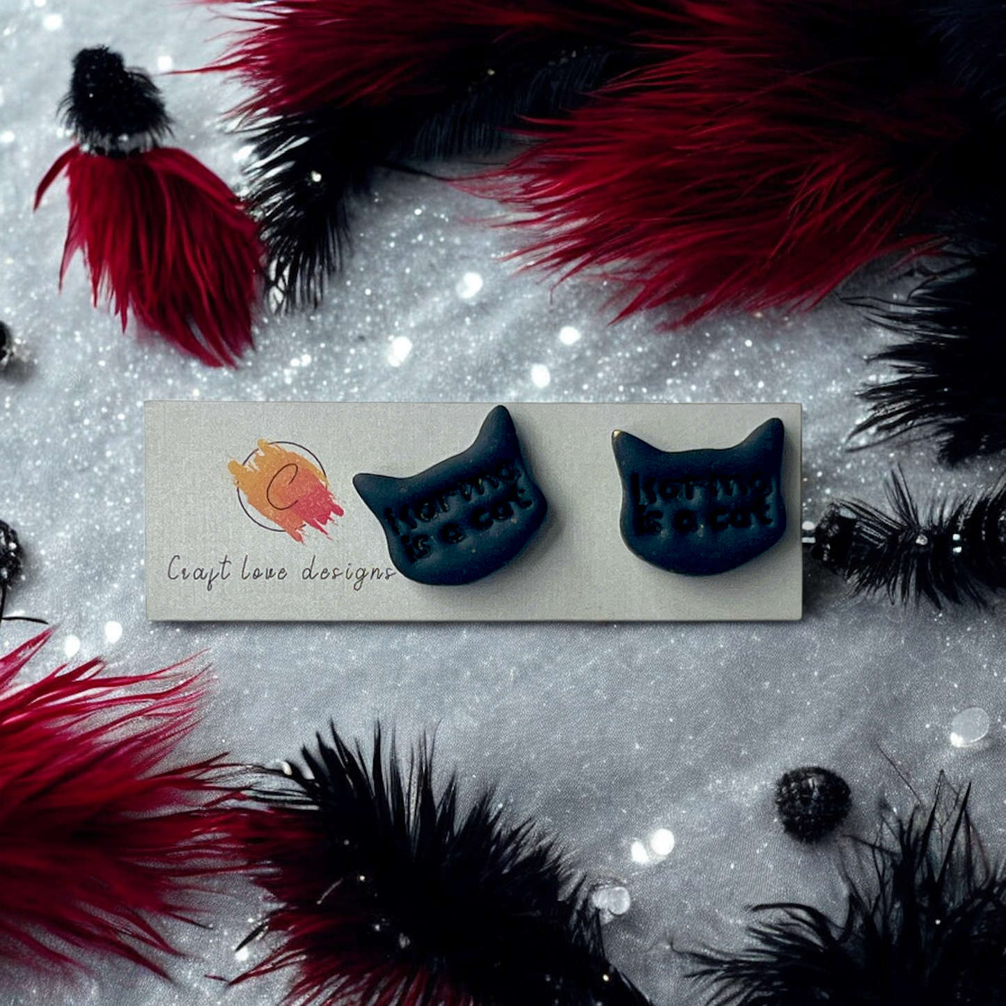 Black cat-shaped stud earrings with embossed text "Karma is a Cat," displayed on a Craft Love Designs card, surrounded by feathers and sparkle.