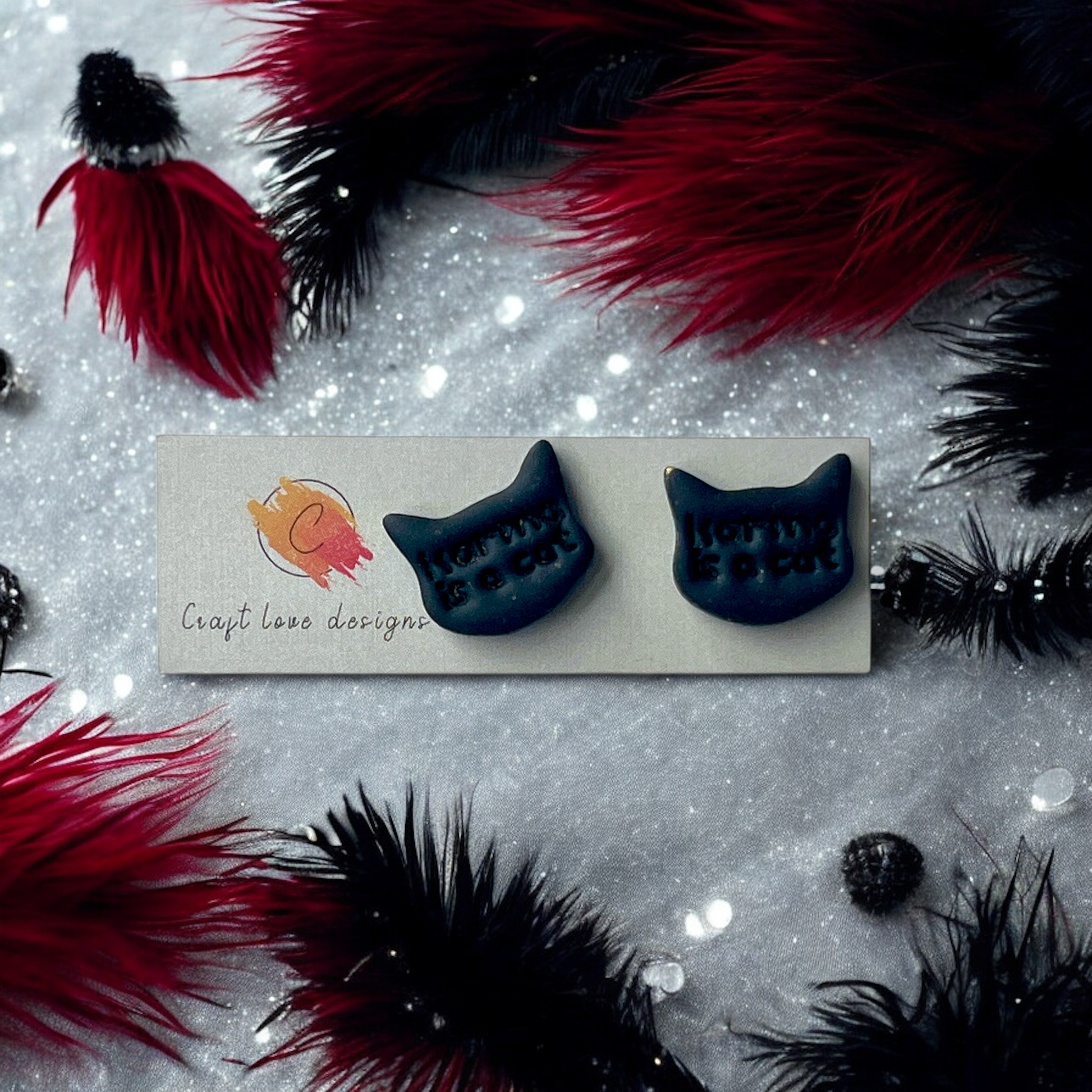 Black cat-shaped stud earrings with embossed text "Karma is a Cat," displayed on a Craft Love Designs card, surrounded by feathers and sparkle.