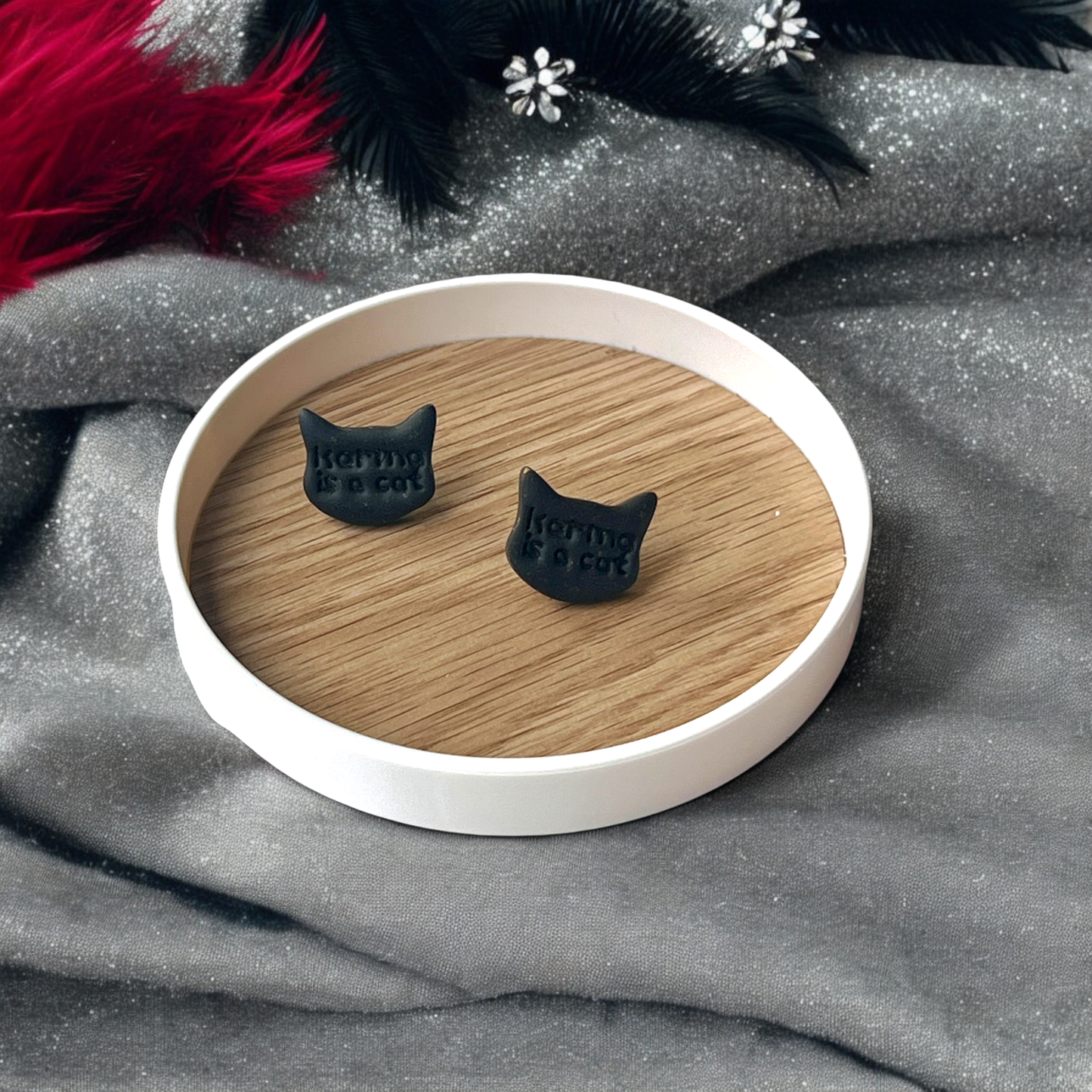 Black cat-themed stud earrings with "Karma is a Cat" text, placed on a wooden display tray, surrounded by soft, silver and red decorative accents.