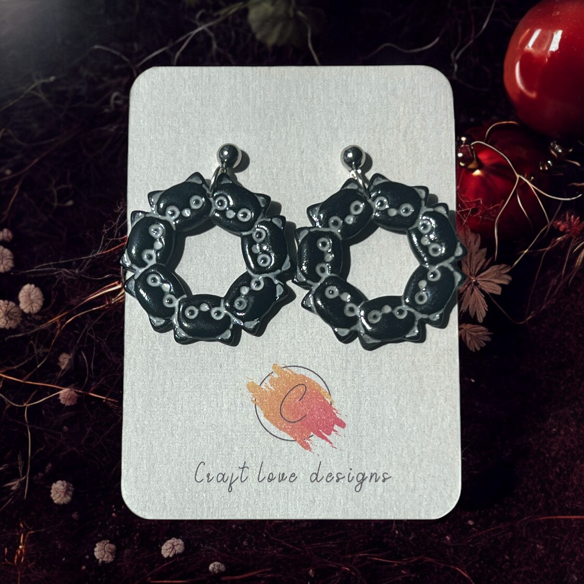 A close-up of black cat-themed wreath earrings on a Craft Love Designs card, surrounded by dark, moody decor.