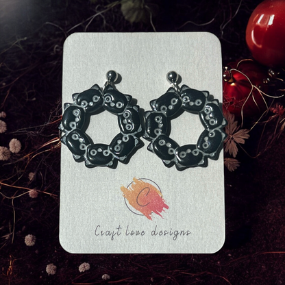 A close-up of black cat-themed wreath earrings on a Craft Love Designs card, surrounded by dark, moody decor.