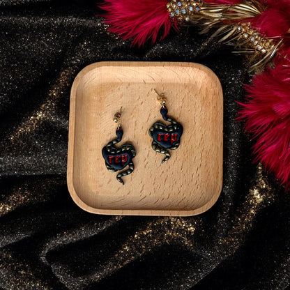 Pair of black heart earrings featuring coiled gold snakes and red “rep” text, resting on a wooden tray against a glittery black and red setting.