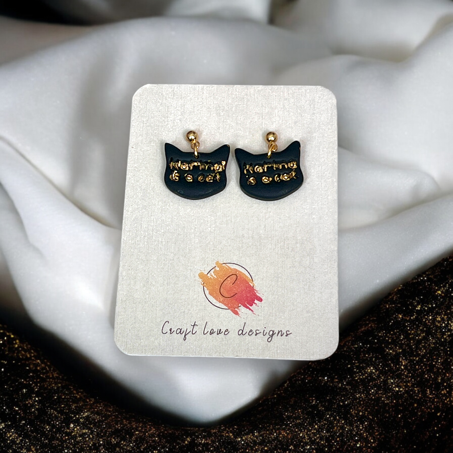 Black cat-shaped dangling stud earrings with gold-embossed "Karma is a Cat" text, displayed on a Craft Love Designs backing card.
