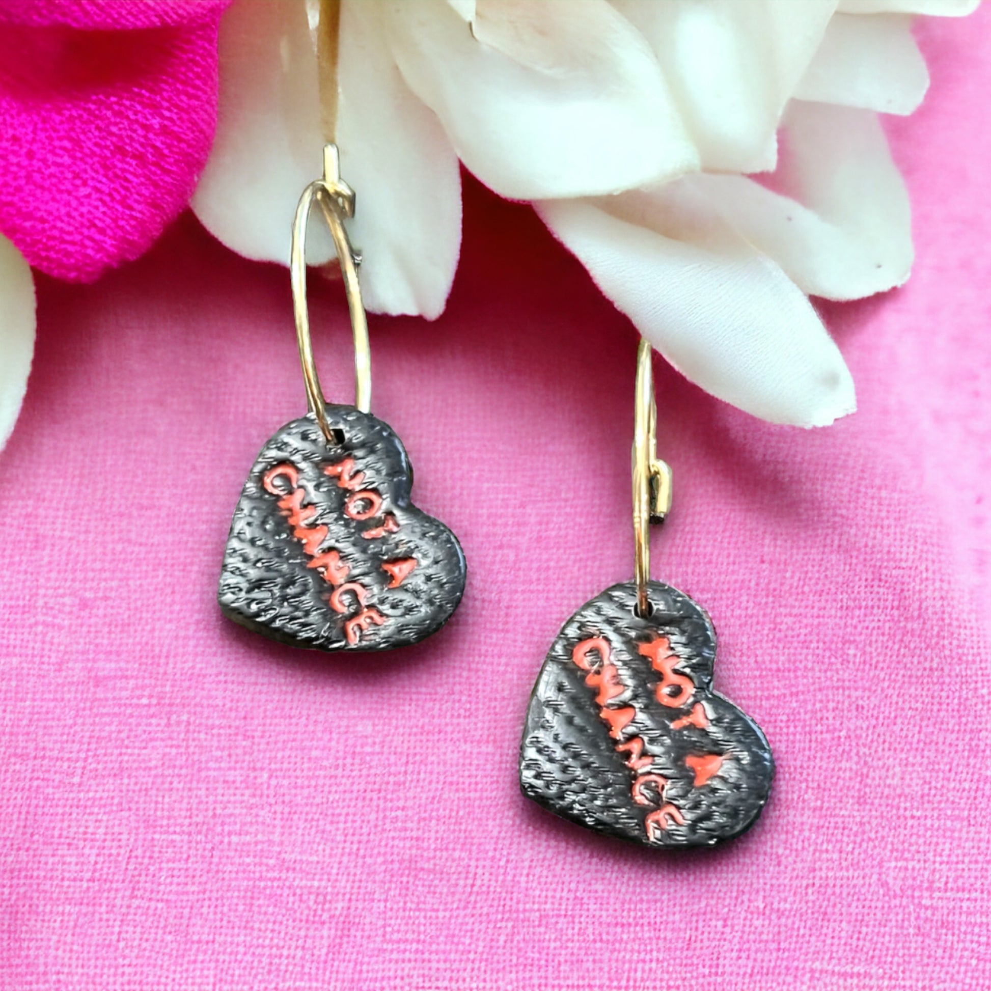 Close-up of black heart-shaped hoop earrings featuring “Not A Chance” text in red on a textured surface, resting against pink floral decor.