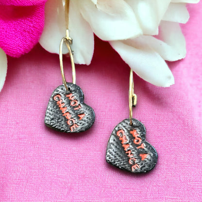Close-up of black heart-shaped hoop earrings featuring “Not A Chance” text in red on a textured surface, resting against pink floral decor.