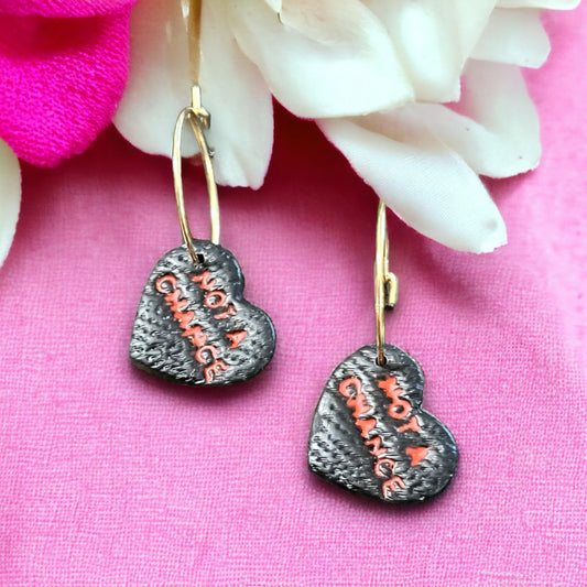 Close-up of black heart-shaped hoop earrings featuring “Not A Chance” text in red on a textured surface, resting against pink floral decor.