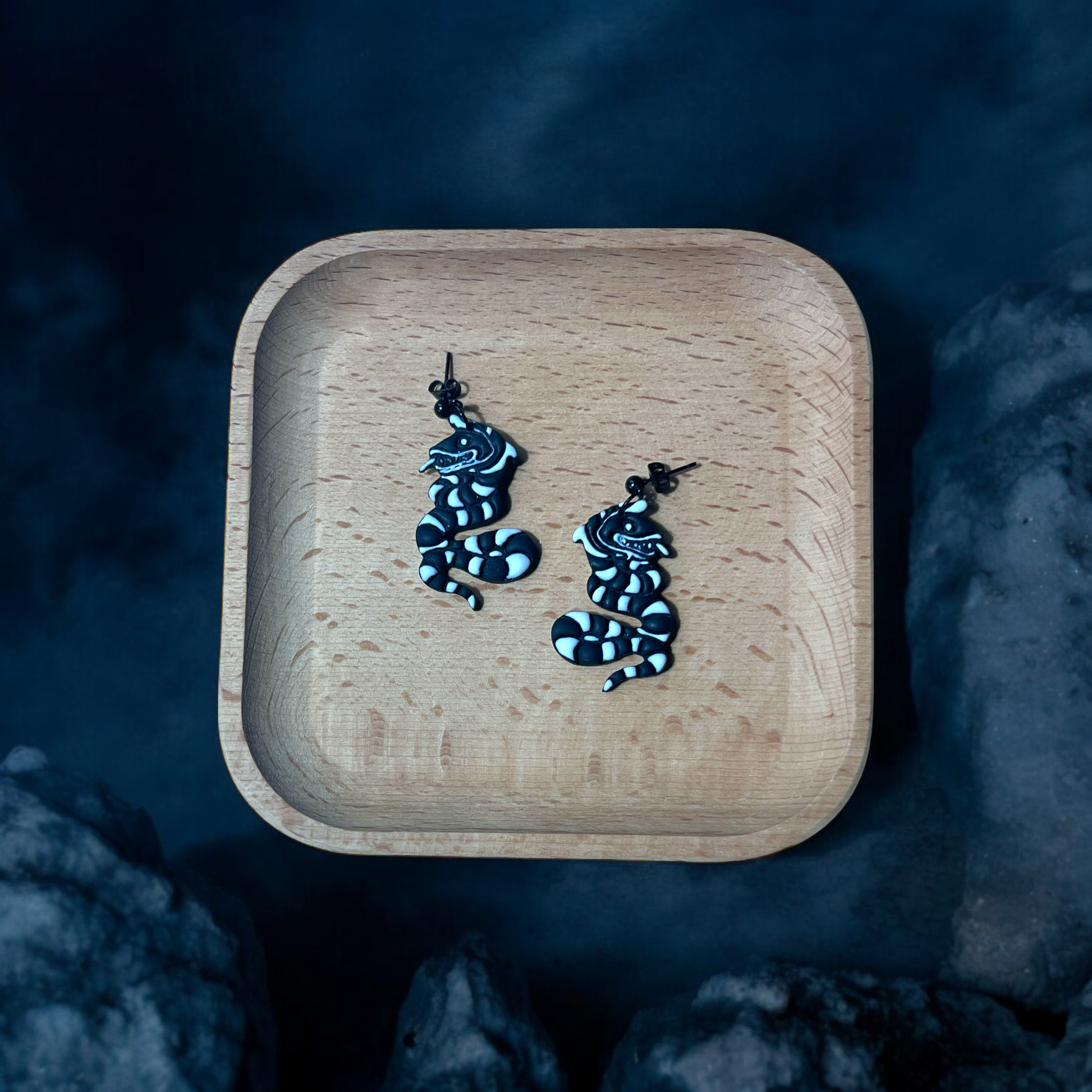 Black striped snake earrings placed in a wooden tray with a smoky, eerie backdrop, creating a mysterious and Halloween-themed display. 