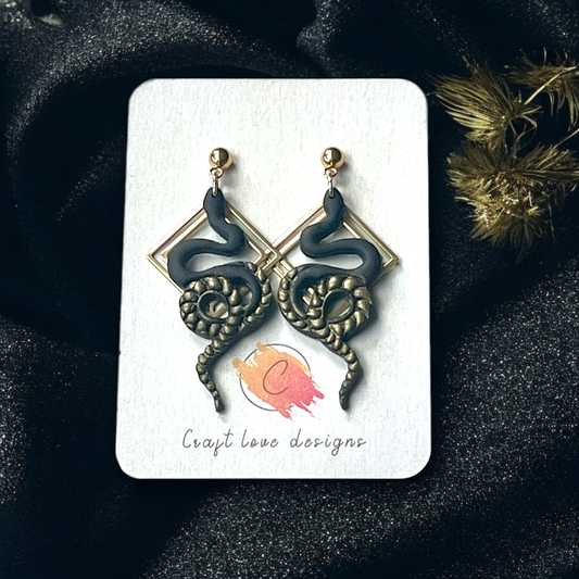 Black snake earrings coiled around geometric gold frames with golden stud tops, displayed on a branded card against dark fabric. 