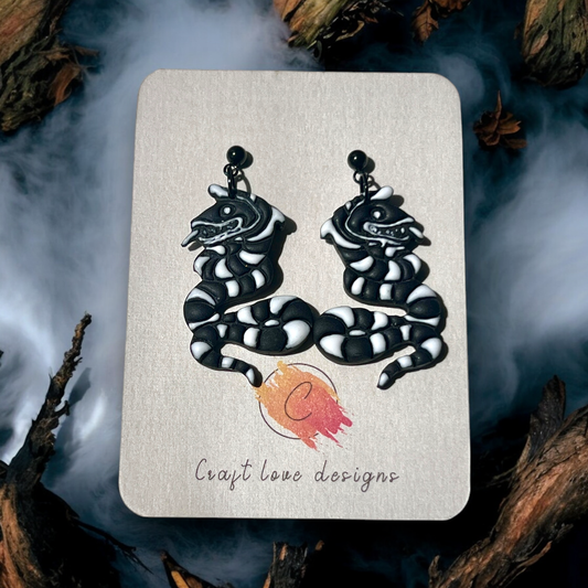 Black striped snake polymer clay earrings on Craft Love Designs display card, styled with a smoky background for a spooky, mystical vibe. 