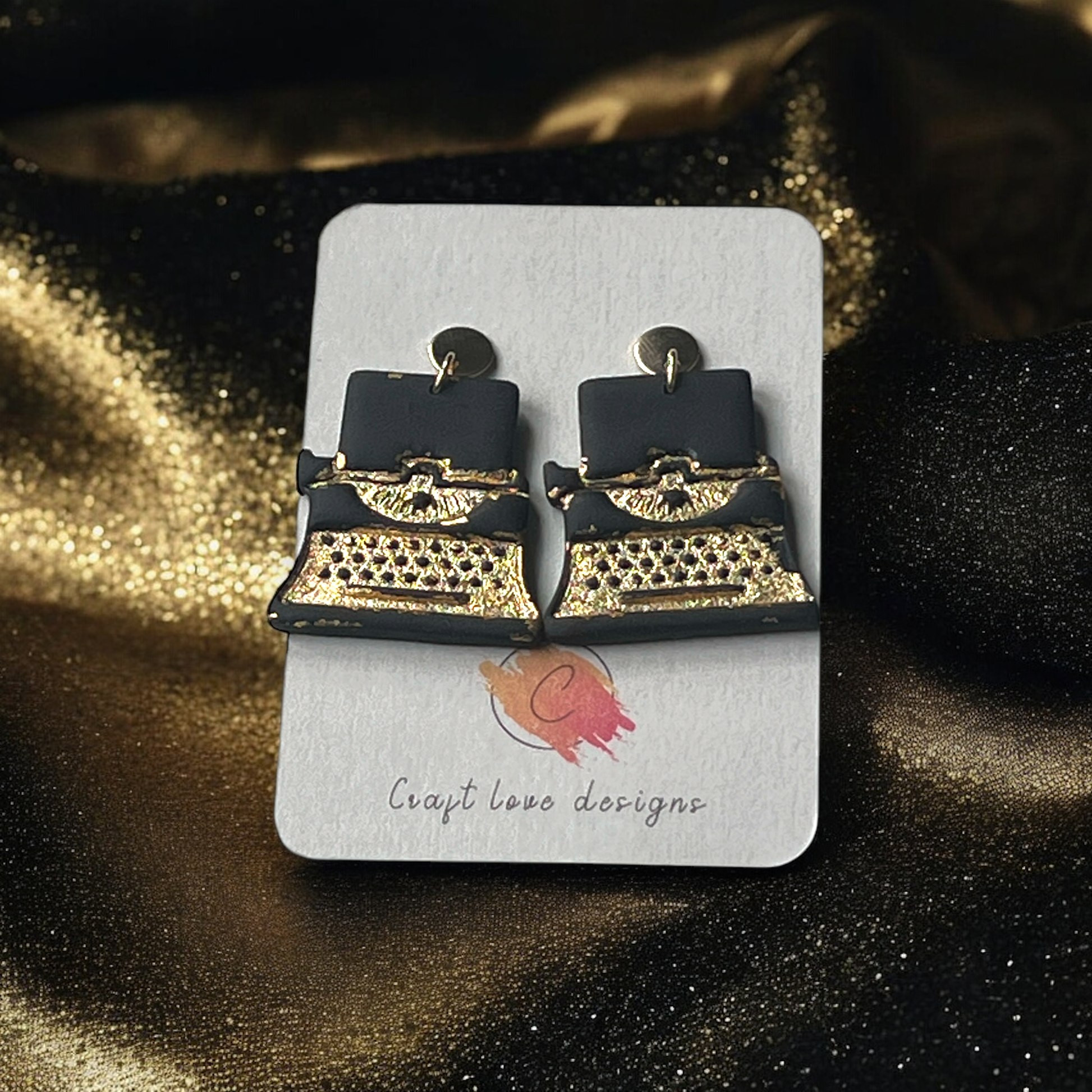 Black and gold typewriter-inspired earrings displayed on a Craft Love Designs card, set against a shimmering gold and black background.