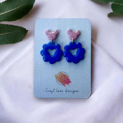 Cute blue flower-shaped earrings with a heart cutout and pink stud tops displayed on a card, placed on soft white fabric with green leaves. 