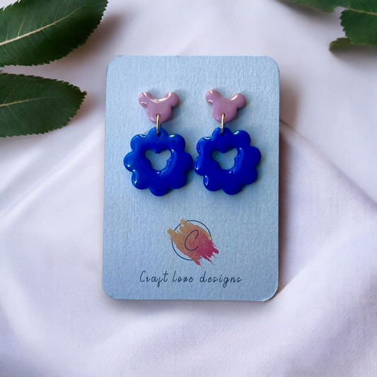 Cute blue flower-shaped earrings with a heart cutout and pink stud tops displayed on a card, placed on soft white fabric with green leaves. 