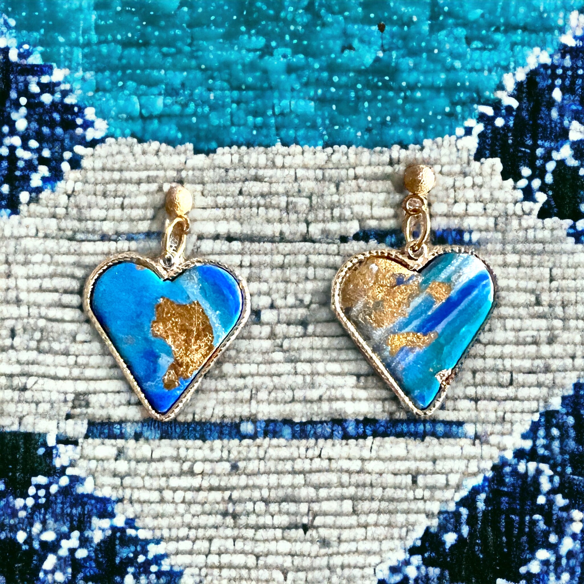 Close-up of elegant blue heart-shaped dangle earrings with gold accents, placed on a textured blue rug background.