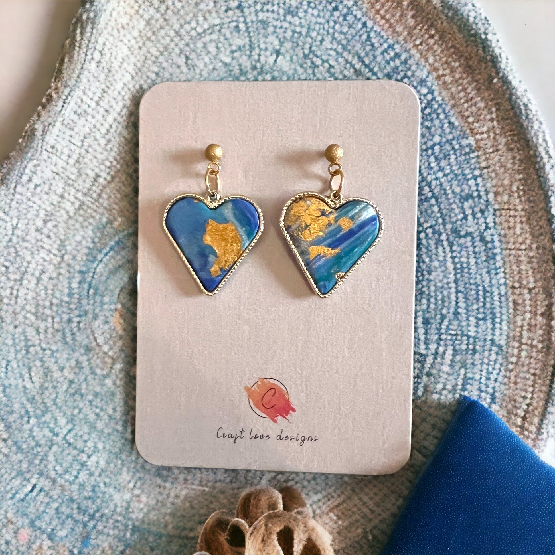 A pair of heart-shaped dangle earrings in blue and gold displayed on a product card with artistic textures in the background. 