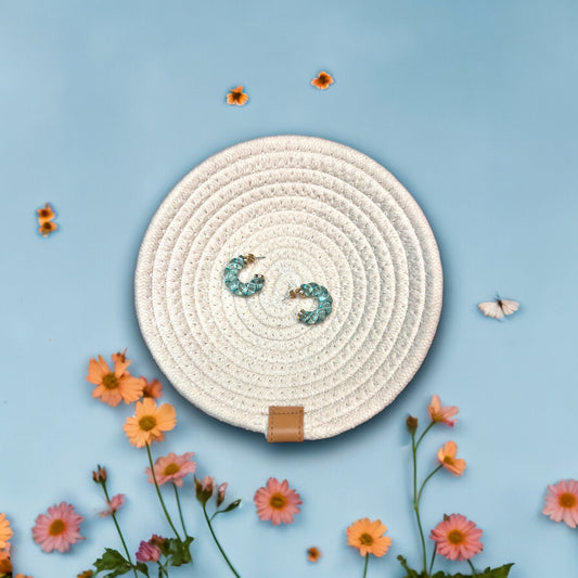 A pair of Blue Summer Braided C-Hoop Earrings displayed on a woven round white mat, surrounded by colorful flowers and butterflies on a sky-blue backdrop.