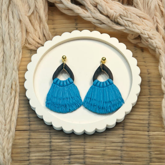 Close-up of handmade blue fringe drop earrings with a dark wooden frame and gold accents, displayed on a white decorative plate.