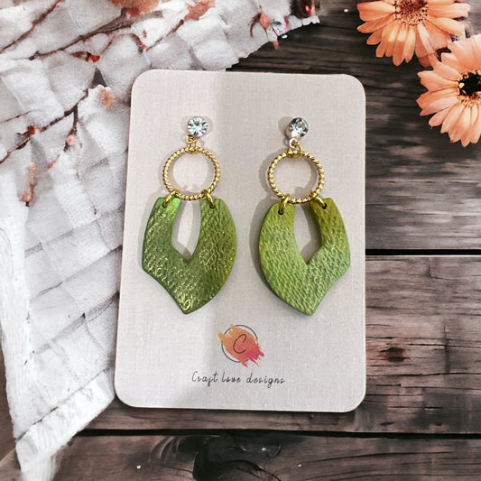 Green boho dangle earrings with textured leaf-shaped pendants, gold hoop details, and cubic zirconia studs on a wooden backdrop. 