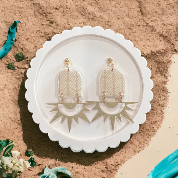 Elegant clear acrylic dangle earrings with sunburst and chevron patterns, displayed on a white scalloped plate against a sandy background.