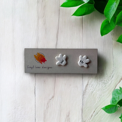 White cartoon-style hand stud earrings displayed on a designer card placed on a light wooden surface with green leaves in the background. 