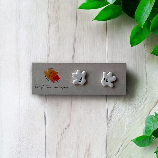 White cartoon-style hand stud earrings displayed on a designer card placed on a light wooden surface with green leaves in the background. 
