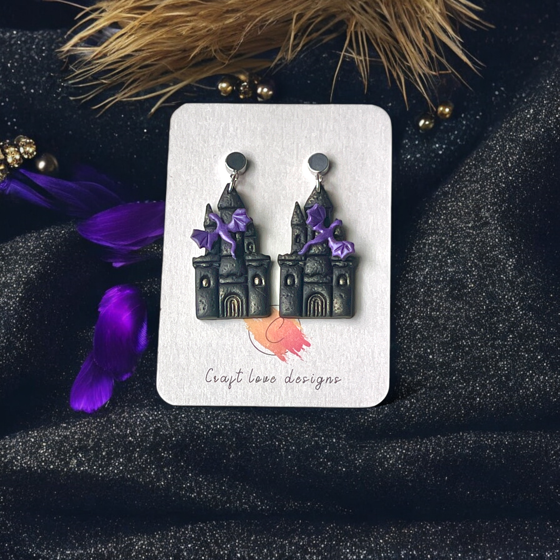 Gothic castle-themed earrings with purple bat accents displayed on a Craft Love Designs card, set against a dark, sparkly background.