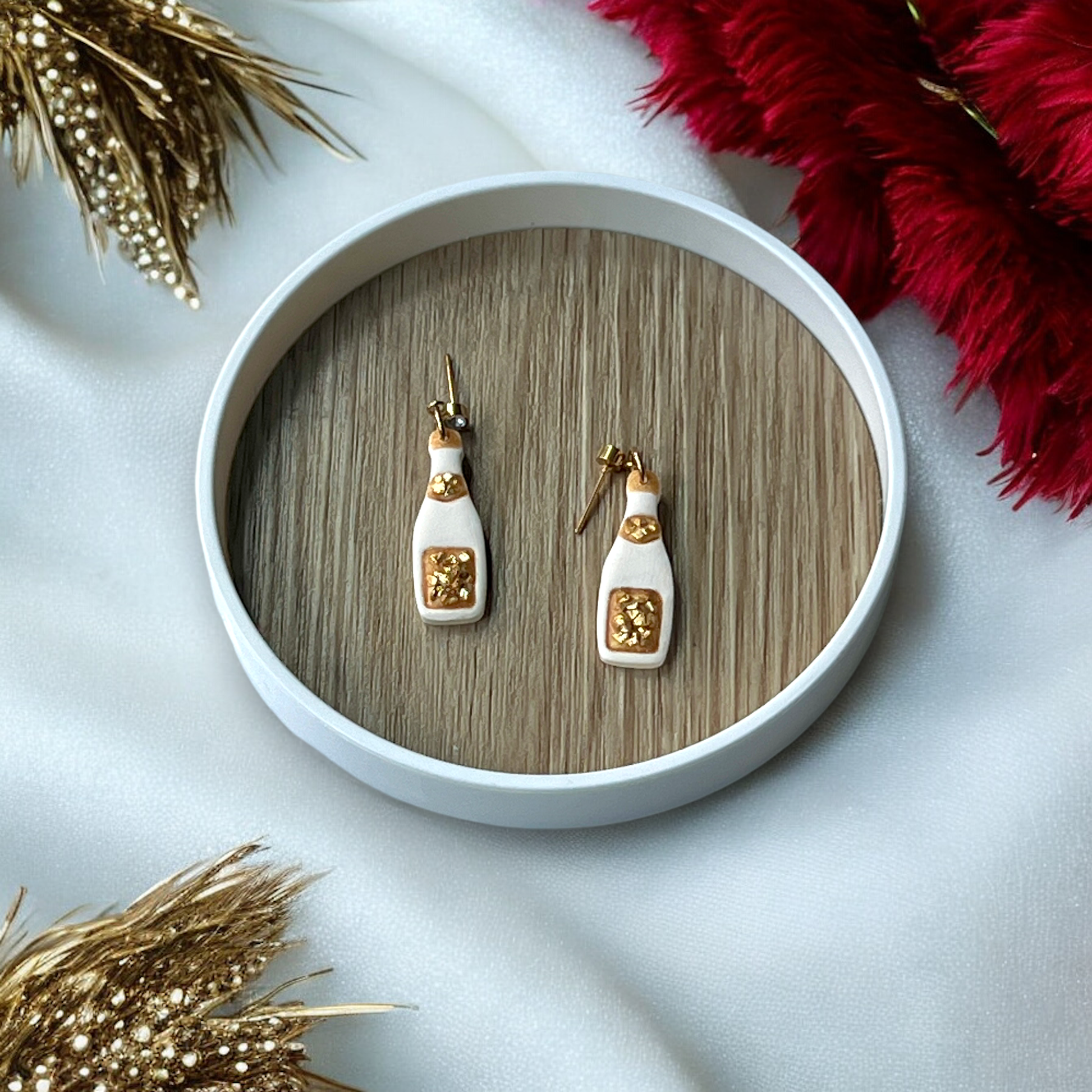 Gold-accented champagne bottle earrings resting in a round wooden tray, surrounded by white satin, red feathers, and sparkling décor.