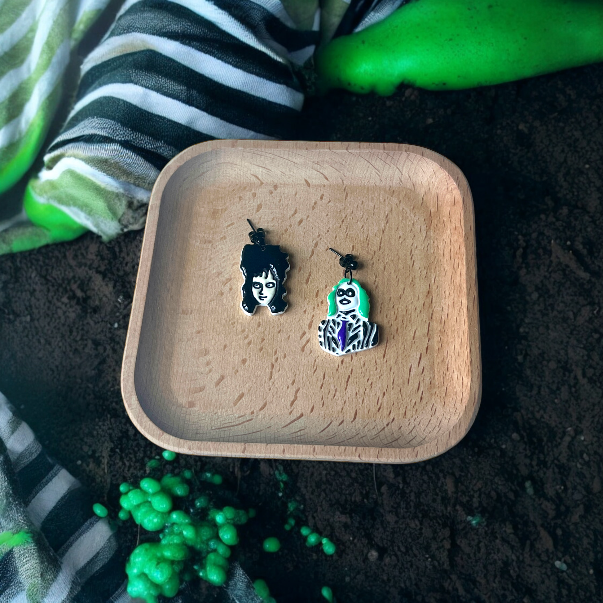 Black and green-haired character-inspired earrings resting in a wooden tray, surrounded by striped fabric and green spooky accents. 