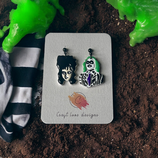 Character-inspired earrings featuring black hair and green-haired figures on a Craft Love Designs card, set against dirt and spooky decor. 